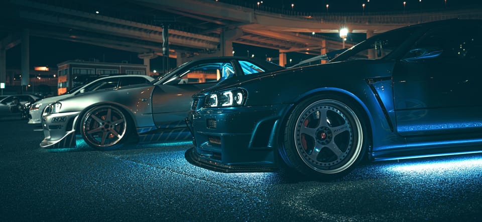 Nissan Skyline R34 GT-Rs with neon underglow lined up at a Daikoku PA night car meet under expressway overpasses