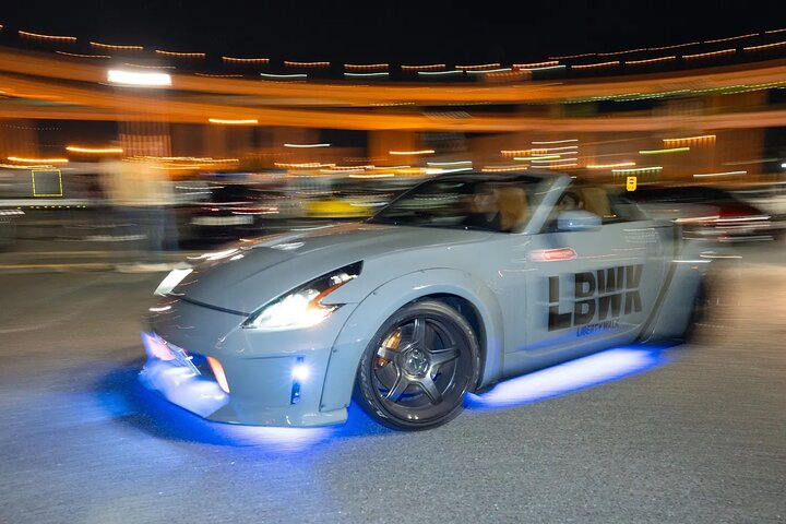 Liberty Walk Nissan 350Z roadster with blue underglow rolling through Daikoku PA at night