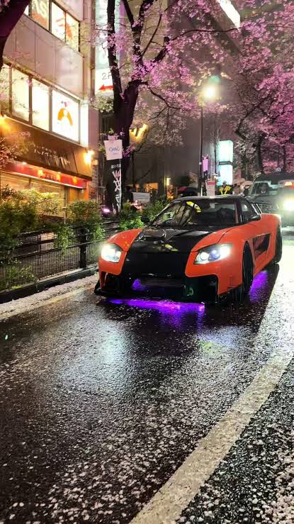 Orange Veilside RX-7 driving under cherry blossoms on a rainy Tokyo street at night